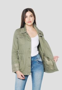 Olivno zelena bombažna jakna z velikimi žepi, okrasnim šivom na rokavih in zapiranjem na gumbe. Nošena odprta preko belega topa.