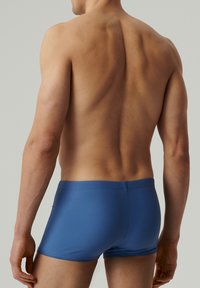 Męskie niebieskie kąpielówki wykonane z gładkiego materiału, charakteryzujące się dopasowanym krojem, niskim stanem i krótkimi nogawkami, prezentujące schludną sylwetkę.