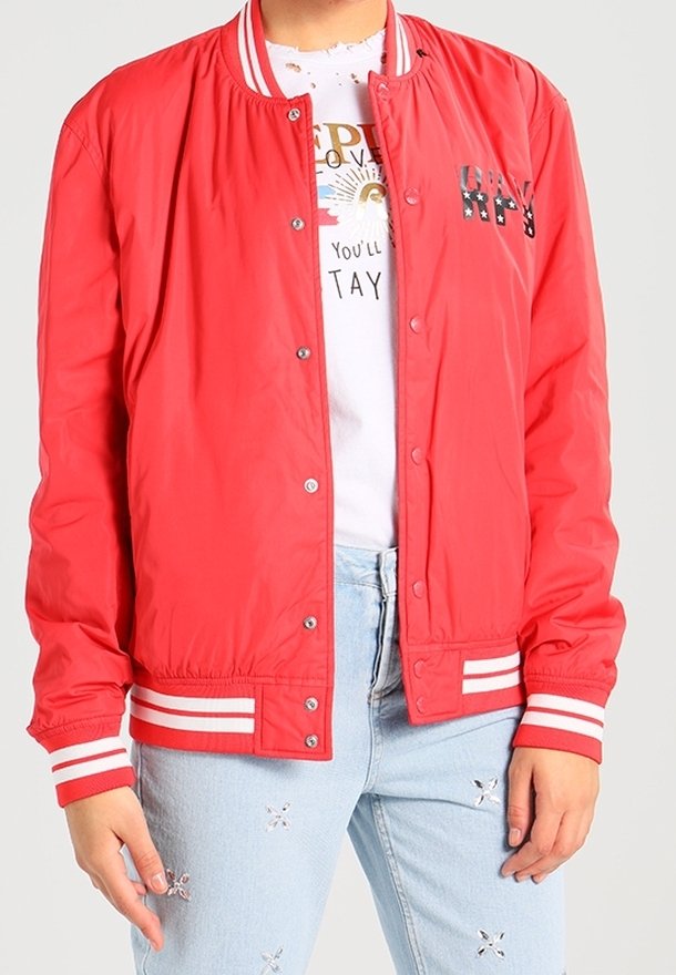 Personne portant une veste rouge boutonnée avec des rayures blanches aux poignets et à l'ourlet, un t-shirt blanc à motif graphique, et un jean bleu clair avec de petites déchirures.
