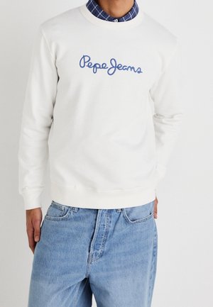 Osoba ubrana w biały sweter z niebieskim haftem "Pepe Jeans", noszony na niebieskiej koszuli w kratę, w połączeniu z jasnoniebieskimi dżinsami.