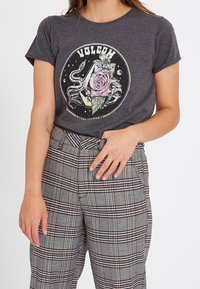 T-shirt en coton gris avec un motif graphique représentant une rose, un serpent et un texte circulaire. Associé à un pantalon taille haute à carreaux noir et blanc.