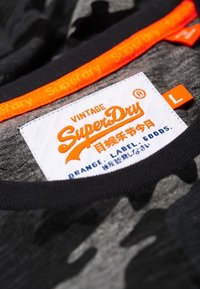 Detailní pohled na šedou kamuflážovou košili SuperDry, na límci je vidět oranžová značka s logem a velikostní štítek "L" na vnitřní straně výstřihu.