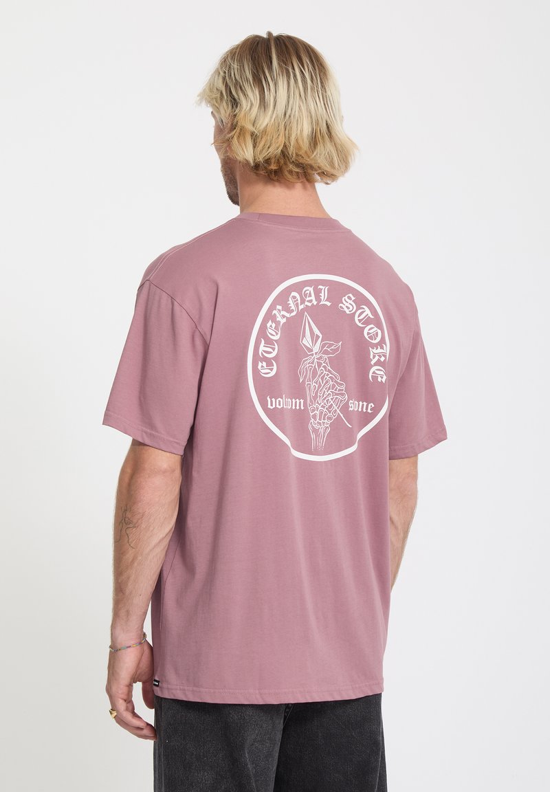 Homme aux cheveux blonds portant un T-shirt mauve avec une main de squelette blanche tenant un cristal et le texte « Pierre Éternelle » au dos.