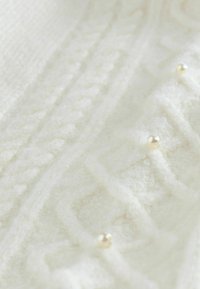 Balts adījuma audums ar tekstūru un maziem pērļu akcentiem, demonstrējot mīkstu, pūkainu virsmu un sarežģītas šuvju detaļas.