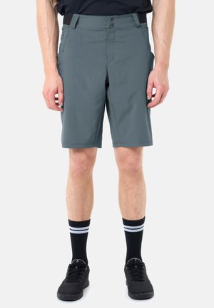 Vista frontale di una persona che indossa pantaloncini grigi fino al ginocchio, scarpe atletiche nere e calzini neri con strisce bianche su sfondo semplice.