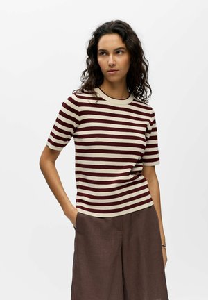 Femme aux cheveux bouclés et foncés portant un haut à manches courtes beige et bordeaux rayé et un pantalon marron à jambes larges, debout avec une main dans la poche.