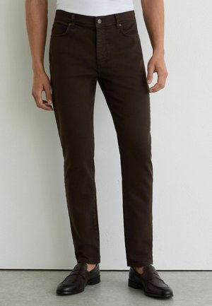 Mand iført slim-fit mørkebrune bukser og sorte læderloafers, stående mod en hvid væg og grå gulv.