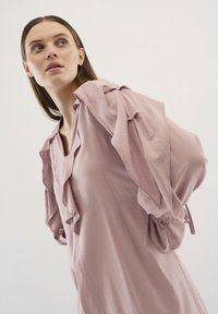 Rochie roz deschis, cu decolteu în V, detalii pliante și mâneci bufante cu accente legate. Material neted cu o textură moale.