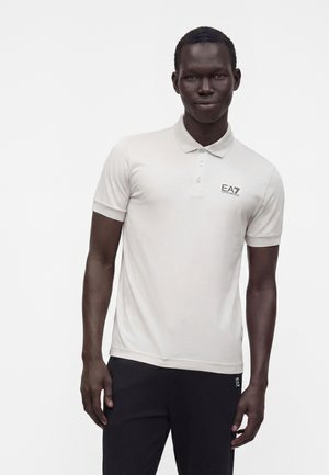 Mann trägt ein weißes kurzärmeliges Polo-Shirt mit EA7-Logo und schwarze Hose und steht vor weißem Hintergrund.