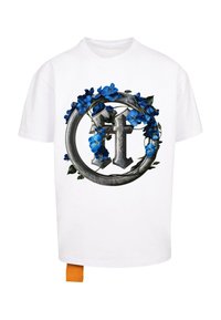 Hvit T-skjorte med et sirkulært design med et dekorativt sølvemblem omgitt av blå blomster og grønne blader. Oransje merkelapp nederst.