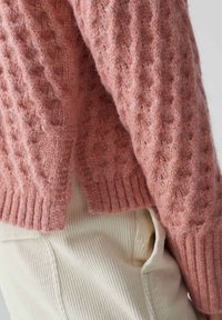 Różowy sweter z dzianiny warkoczowej o fakturowanym wykończeniu i ściągaczu na dole, w połączeniu z kremowymi spodniami z korduroy. Miękka, ciepła tkanina z widocznymi włóknami.