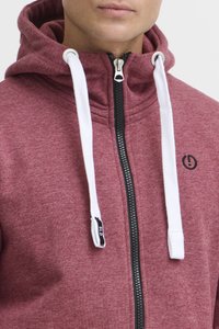 Bordowy hoodie na zamek z wysokim kołnierzem, białymi sznurkami, czarnym zamkiem i małym logo na klatce piersiowej. Miękka faktura materiału.