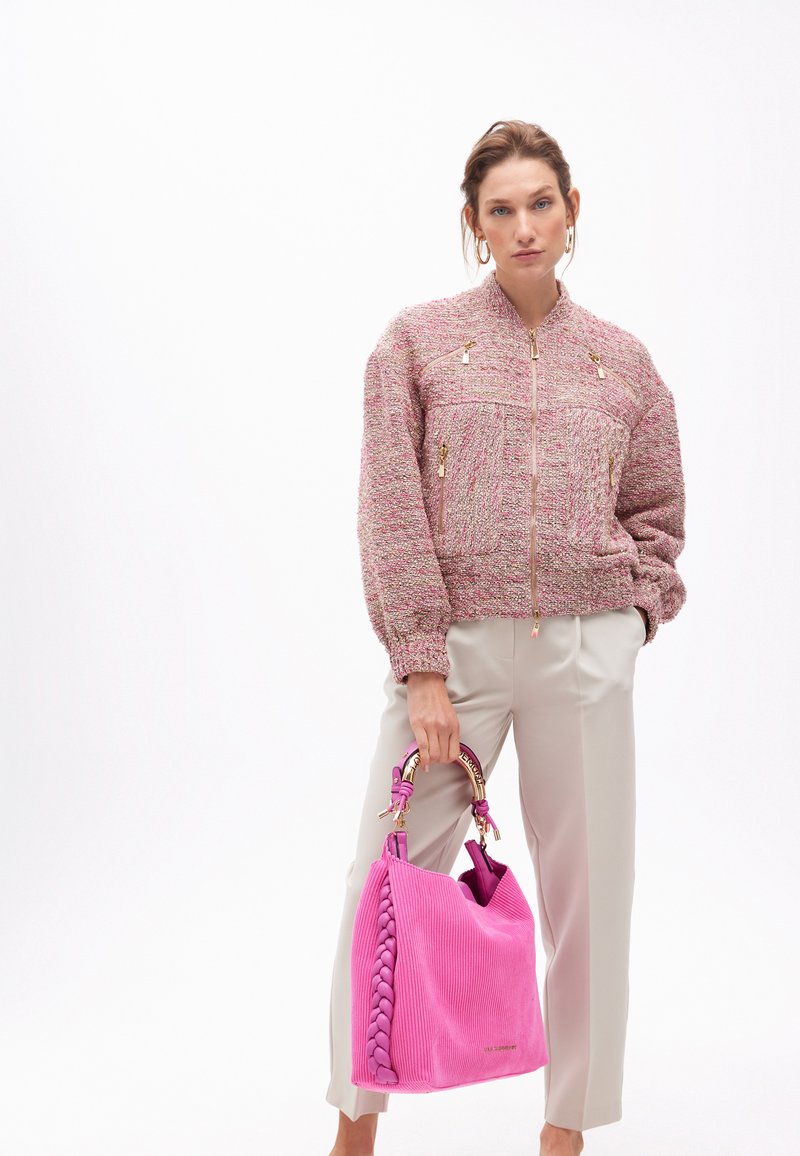Modello in una giacca bomber rosa strutturata e pantaloni crema, che tiene una grande borsa rosa con manici intrecciati su uno sfondo neutro.