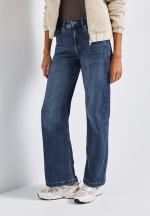 Jean large en denim bleu foncé, avec un design à cinq poches, une fermeture éclair et une légère décoloration. Associé à des baskets claires.