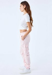 Helle rosa Cargo-Hose mit Seitentaschen und elastischen Bündchen, kombiniert mit einem weißen, kurzen T-Shirt. Das Model steht seitlich und trägt weiße Turnschuhe.