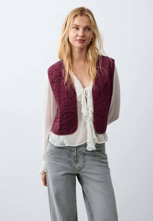 Gesteppte bordeauxrote Weste mit abgerundetem Ausschnitt, getragen über einer weißen, gerafften Bluse, kombiniert mit hellgrauen Jeans mit Verzierungen.