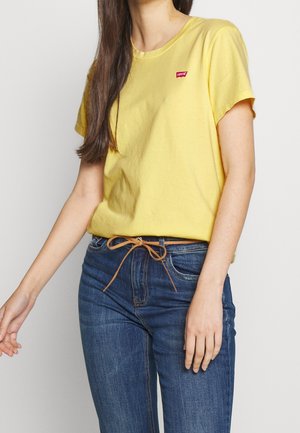 Gul bomuld T-shirt med rund halsudskæring og korte ærmer, med et lille rødt logo mærke. Kombineret med blå højtaljede jeans.