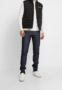 Musta toppaliivi vetoketjulla edessä, päällä kerma villapaita, yhdistetty tummansinisiin farkkuihin ja valkoisiin tennareihin.