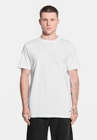 Biała koszulka z krótkim rękawem i okrągłym dekoltem. Wykonana z gładkiej bawełny, charakteryzująca się luźnym krojem. Czarny logo na dolnym brzegu i widoczne tatuaże.