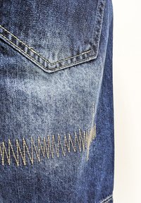Denim jeans in dark blue with a textured finish. Features a zigzag gold stitching pattern along the thigh and a pocket with a pointed flap.