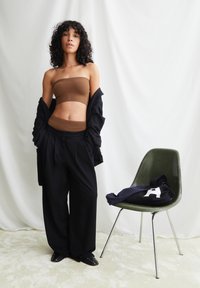 Bruin strapless top gecombineerd met een zwarte pantalon met hoge taille. Model staat naast een groene stoel met een marineblauwe top erop gedrapeerd. Neutrale achtergrond.
