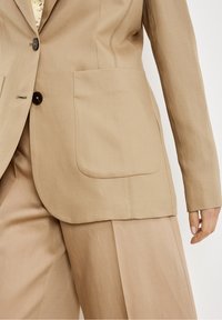 Blazer beige avec deux grandes poches avant, fermeture à un seul bouton et texture lisse. Coupe ajustée avec coutures structurées.