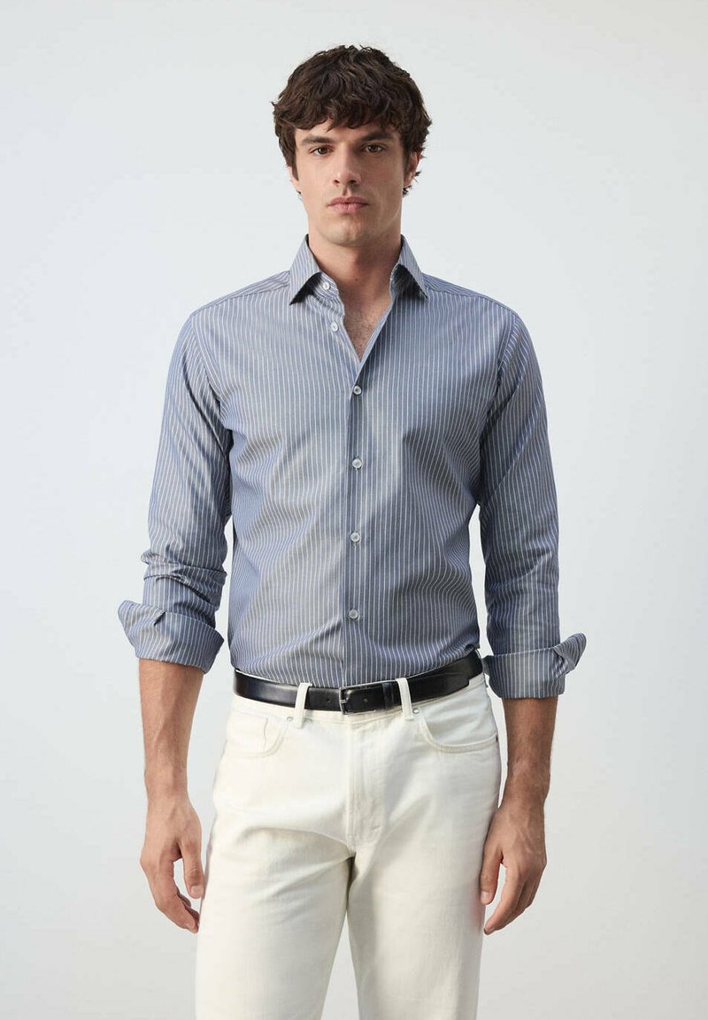 Homme portant une chemise à manches longues rayée bleue avec les manches retroussées, un pantalon blanc et une ceinture noire, debout devant un fond uni.