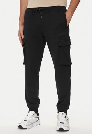 Pantaloni cargo neri in cotone felpato con vita elastica e cordoncino, dotati di tasche laterali e discreto dettaglio del logo. Accoppiati con sneakers bianche.