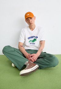 T-shirt blanche avec un design graphique bleu et vert, pantalon large vert, et baskets marron. Casquette orange. Assis sur une surface verte.