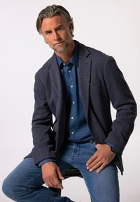 Granatowa teksturowana marynarka nałożona na dżinsową koszulę, uzupełniona niebieskimi dżinsami. Zauważalne detale to chusteczka w kieszeni oraz zegarek na nadgarstku.