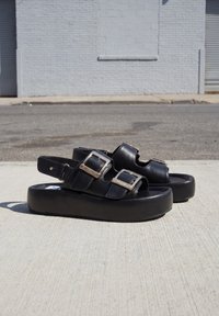 Sandales à plateforme noires avec deux boucles argentées sur chaque, posées sur un trottoir en béton près d’un mur en briques grises et d'une rue vide.