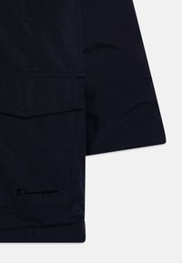 Chaqueta azul marino con una textura suave, que cuenta con un gran bolsillo frontal y un sutil logo de Champion. Diseño liso sin patrones adicionales.