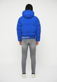 Kék puffertakaru dzseki kapucnival és bordás aljjal. Illeszkedő szürke farmernadrággal és fehér cipővel párosítva. Az Armani logó látható a dzsekiben.