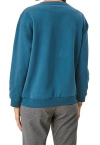 Felpa oversize color teal con colletto rotondo, polsini e orlo ribattuti. Indossata con pantaloni grigi. Tessuto morbido.