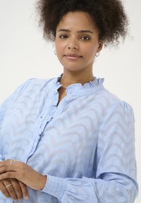 Blouse bleu clair avec un motif en zigzag, dotée d'un col froncé, d'une fermeture à boutons et de manches longues bouffantes avec des poignets élastiques.