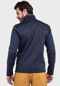 Navy-Fleece-Jacke mit hohem Kragen, strukturiertem Muster und elastischen Bündchen. Stylisch kombiniert mit goldenen Hosen, die ein tailliertes Rückendesign präsentieren.