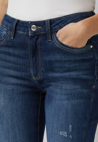 Ciemnoniebieskie dżinsy z wysokim stanem, srebrnym guzikiem i przednimi kieszeniami. Materiał ma delikatne przetarcie w okolicy kolana.