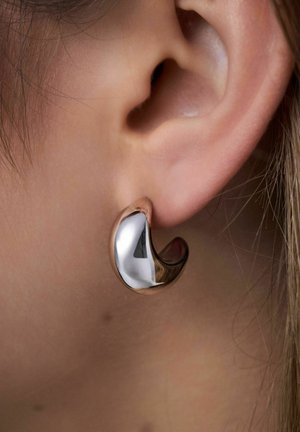 Metallisk ringörhänge med en polerad silveryta och en böjd roséguldaccent. Slät textur, medelstorlek, sitter tätt mot örsnibben.