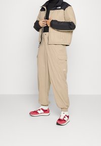 Beige isolerad jacka med svart topp, beige cargo-byxor och röda New Balance sneakers. Funktioner inkluderar elastiska manschetter och en logotyp på jackan.