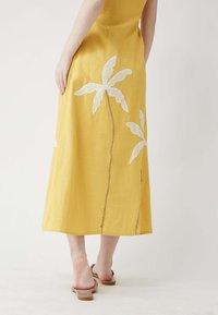 Gelbes Leinenkleid mit weißen gestickten Palmeblättern und geradem Saum. Das Modell trägt sandfarbene Sandalen und zeigt eine entspannte Silhouette.