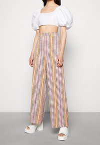 Breda byxor i ett flerfärgat zigzag-mönster med ljusrosa, gult och blått; kombinerat med en vit crop top med puffärmar och platåskor.