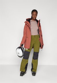 Orange wasserdichte Jacke, gestrickter pinker Pullover, grüne und schwarze Skihose, schwarze Skischuhe und ein schwarz-weißer Helm.