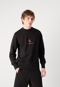 Felpa nera in misto cotone, con scollo rotondo, polsini a coste e un logo ricamato arancione sul petto.