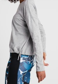 Grå långärmad topp med rundad nederkant och dragsko vid midjan, med vit "new balance" logotyp på ärmen, ihop med mönstrade leggins.