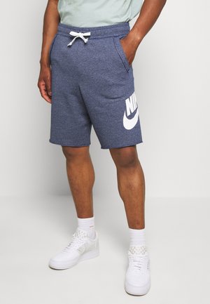 Mężczyzna w niebieskich szortach Nike z białym sznurkiem, białych skarpetkach i białych trampkach, stoi na tle jednolitego szarego tła.