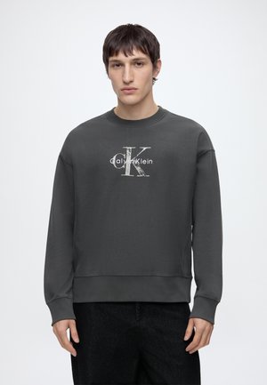 Junger Mann trägt einen dunkelgrauen Calvin Klein Sweatshirt mit Logo auf der Vorderseite und steht vor einem schlichten weißen Hintergrund.