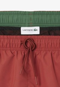Pantaloni da bagno rossi con vita elastica verde. Presentano un cordoncino di regolazione e un'etichetta con il logo Lacoste. Texture liscia e fodera in rete traspirante.