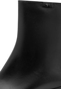 Bottine en cuir noir avec une texture lisse et une petite décoration croix argentée près du bord supérieur.