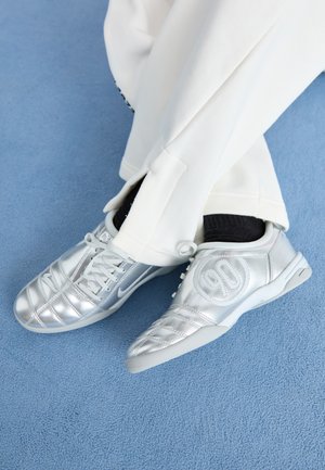 Silver athletic shoes with a smooth, shiny finish, featuring a stitched design and the number 90 on the side, paired with white pants.