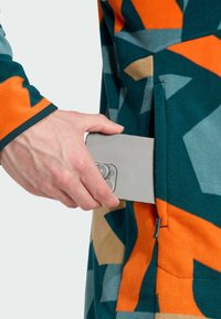 Fleecejacke mit geometrischem Muster in Orange, Türkis und Braun, ausgestattet mit einer Seitentasche, während eine Hand sich danach streckt, um ein Smartphone einzuschieben.